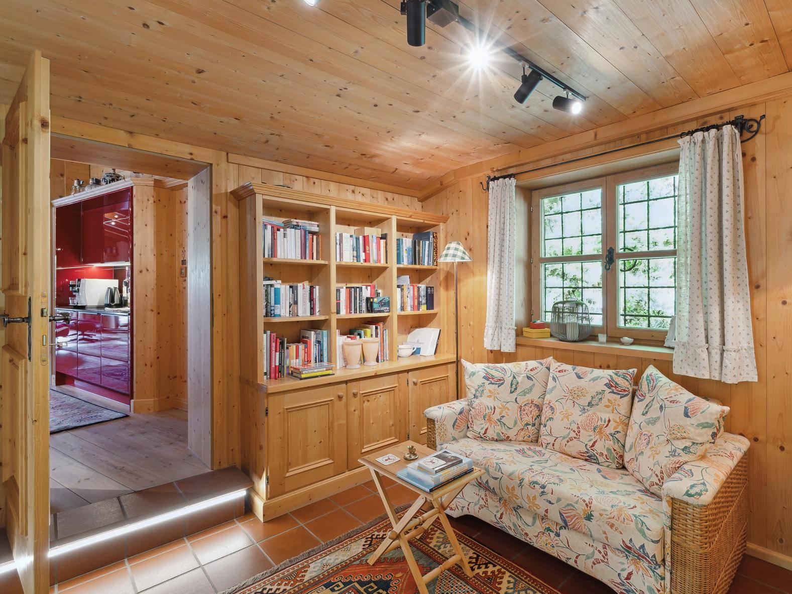 Heller Wohnbereich mit Holztäfelung, Bücherregal und Blick auf eine moderne rote Küche im Hintergrund.