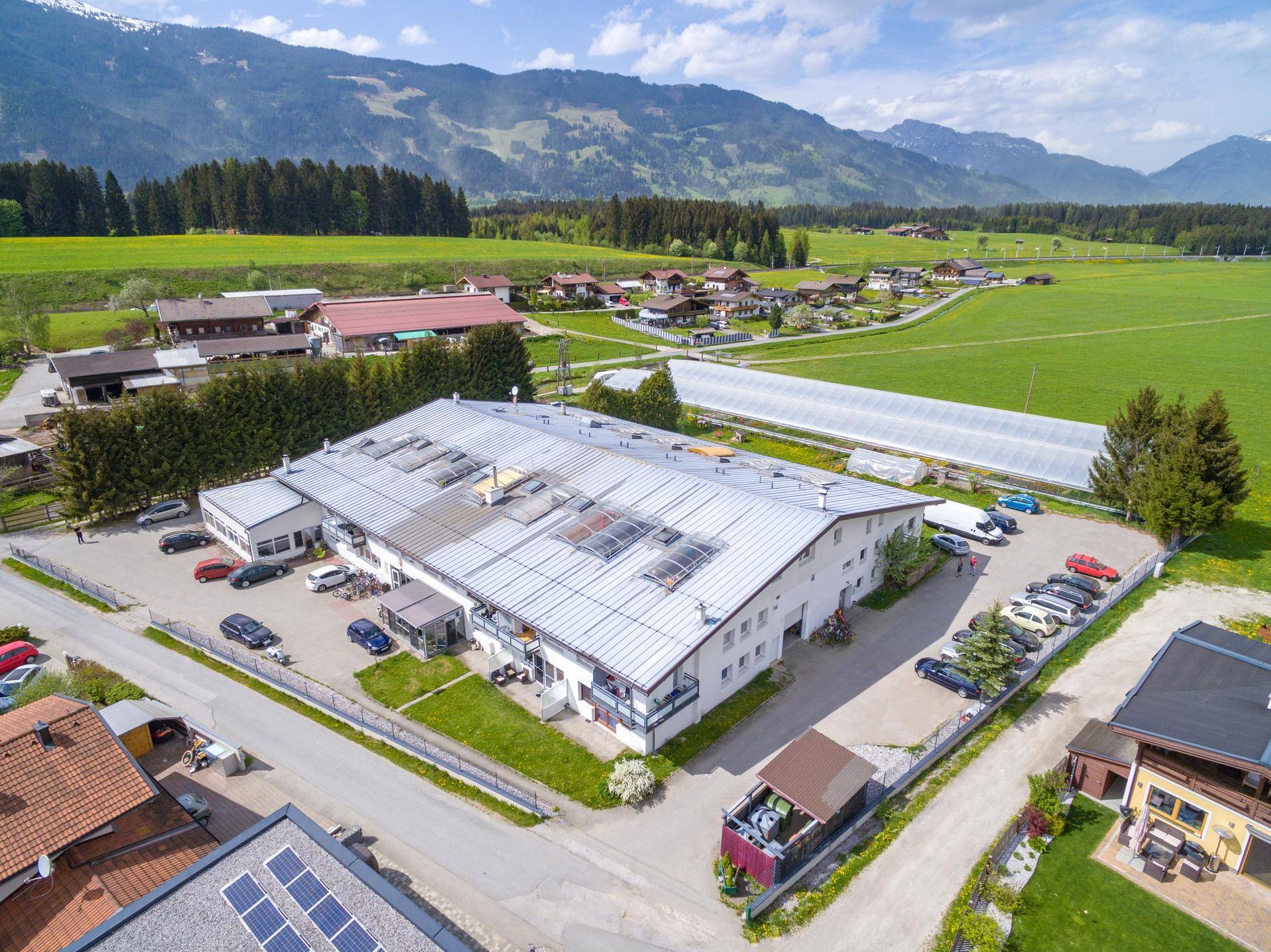 Weitläufige Luftaufnahme eines Gebäudes mit Parkplätzen, umgeben von grünen Feldern und Bergen.