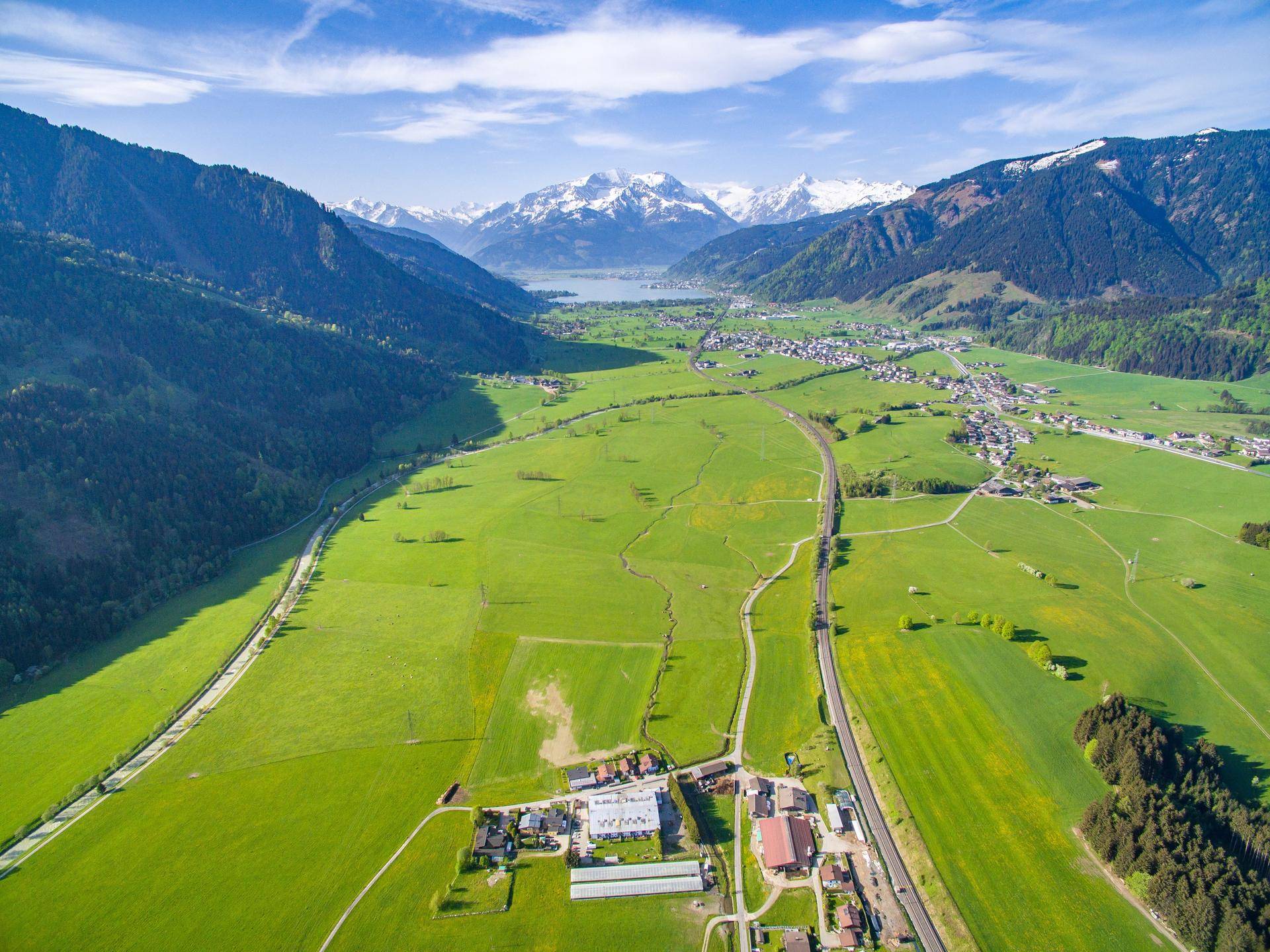 Panoramablick über ein grünes Tal mit See, schneebedeckten Bergen und kleinen Siedlungen.