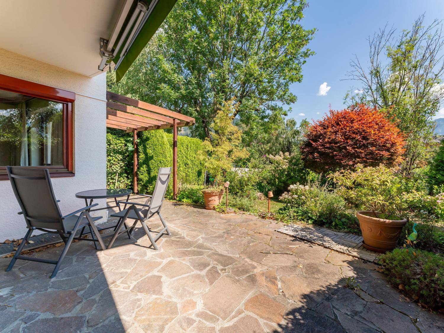 Gemütliche Steinterrasse mit Gartenmöbeln und einer Holzpergola, umgeben von üppiger Vegetation.