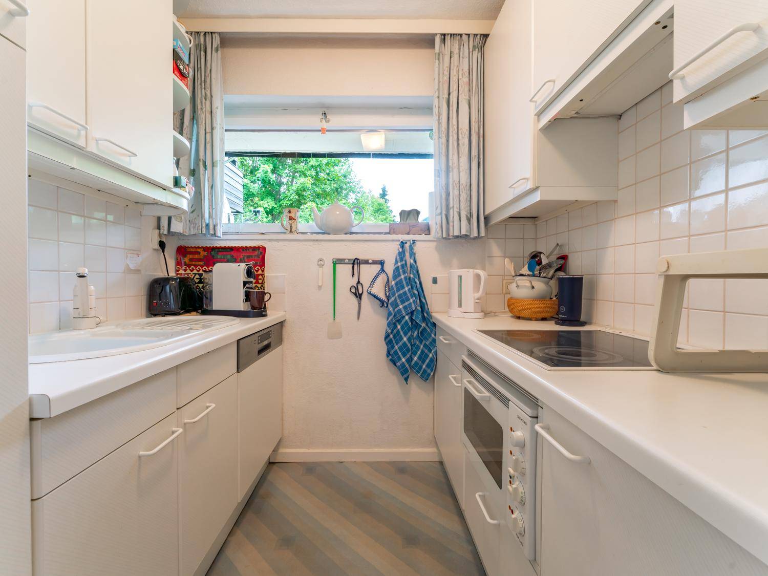 Funktionale, schmale Küche mit weißen Schränken, Fliesenrückwand und einem Fenster für Tageslicht.