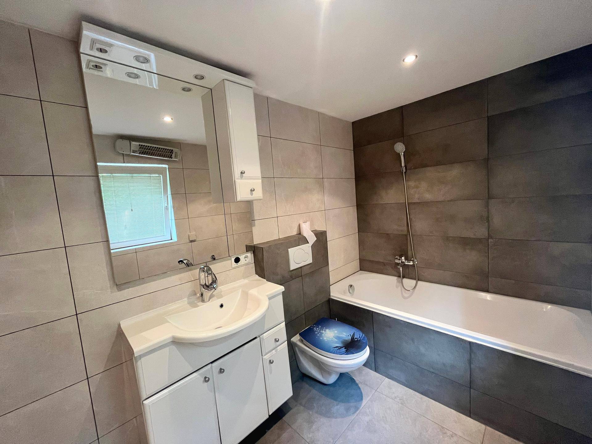 Modernes Badezimmer mit Badewanne, Waschtisch, WC und hellgrauen sowie dunkelgrauen Fliesen.