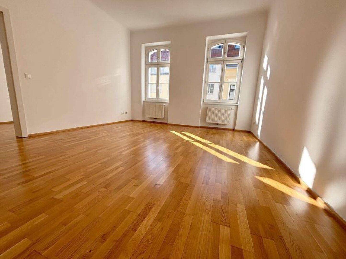 Heller, leerer Raum mit glänzendem Parkettboden und zwei großen Fenstern, die viel Tageslicht hereinlassen.