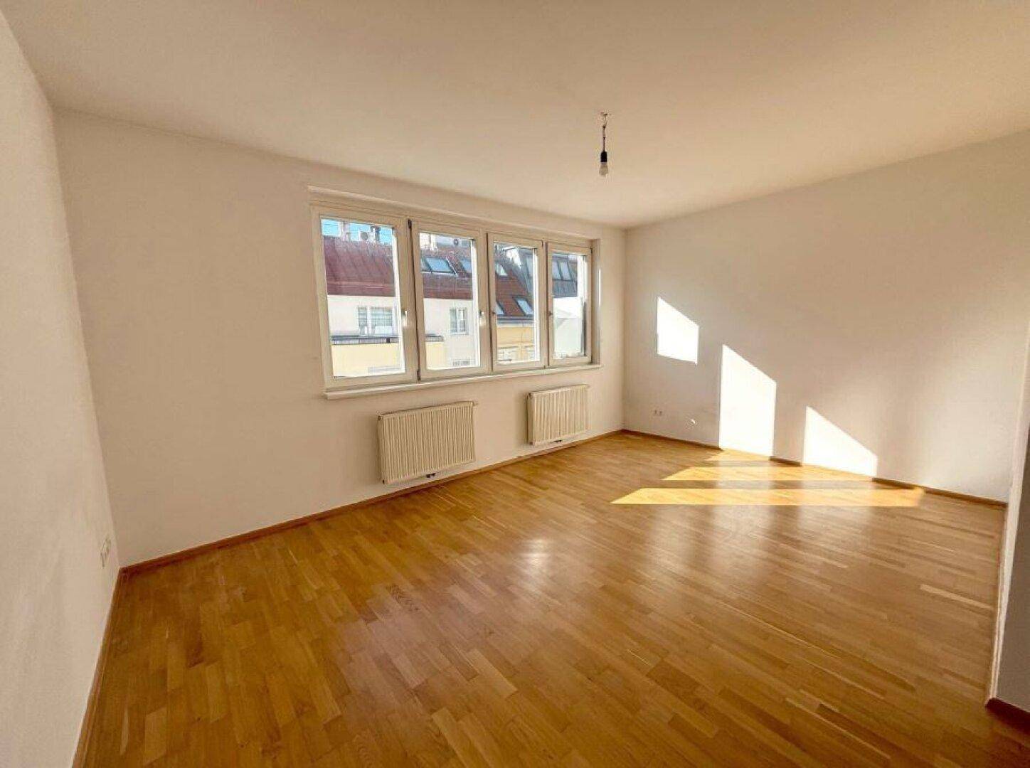 Helles, leeres Zimmer mit Parkettboden und großen Fenstern, die viel Sonnenlicht hereinlassen.