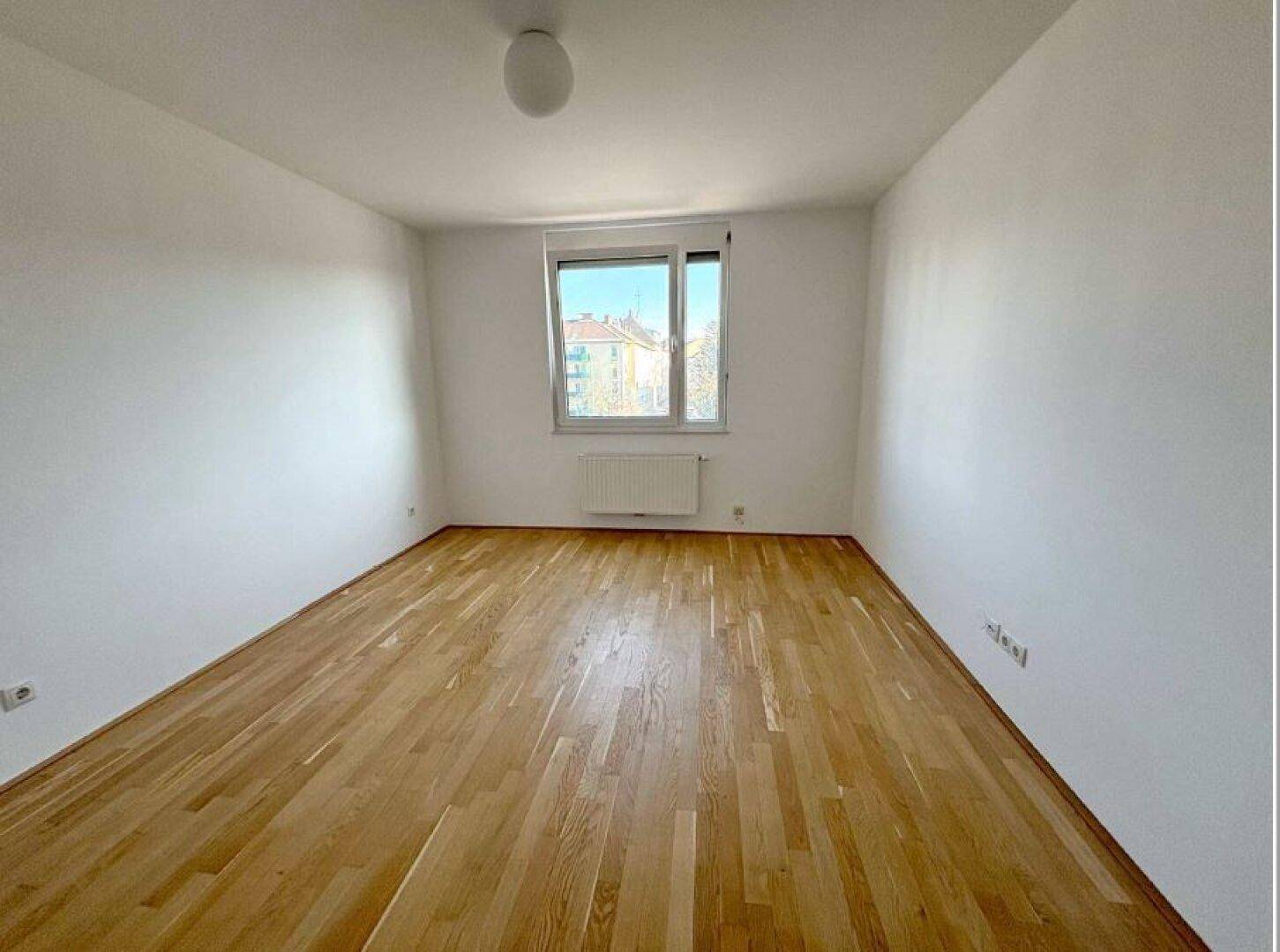 Leeres Zimmer mit hellem Holzparkettboden und einem Fenster, das viel Tageslicht hereinlässt.