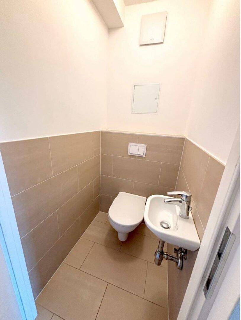 Modernes Gäste-WC mit wandhängender Toilette, kleinem Waschbecken und beigefarbenen Fliesen.