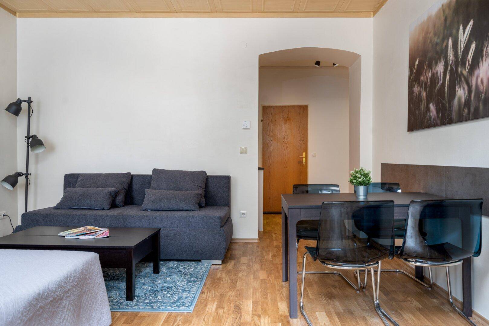 Wohnbereich mit Esstisch, Sofa und Zugang zu einem weiteren Raum durch einen Bogen.