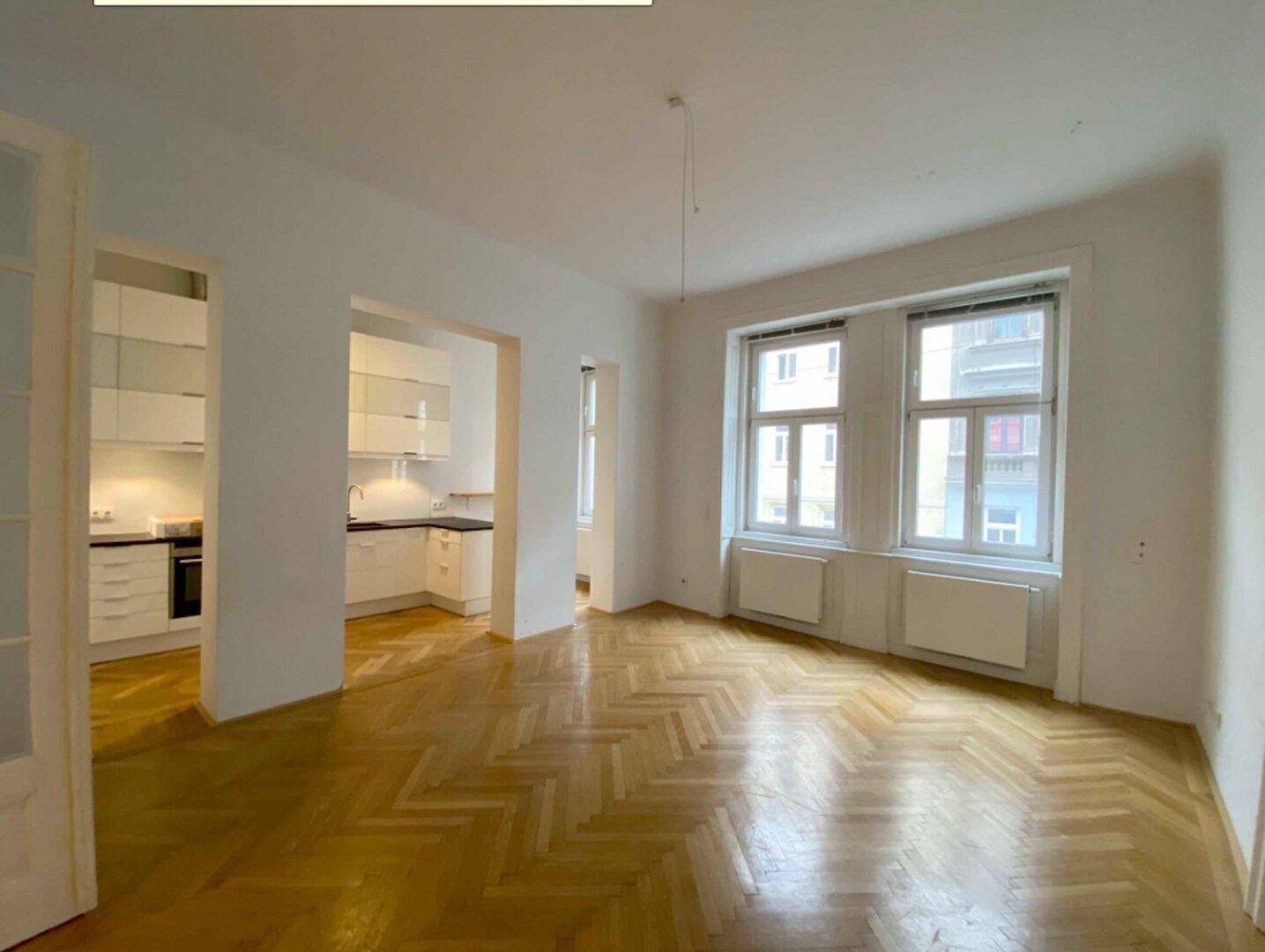 Geräumiges Wohnzimmer mit Fischgrätparkett und Blick in eine angrenzende Küche.