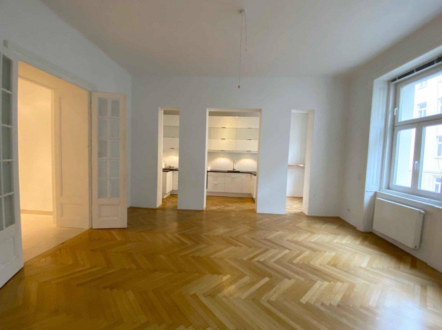 Großes Wohnzimmer mit Fischgrätparkett, hohen Decken und Zugang zu einer modernen Küche.