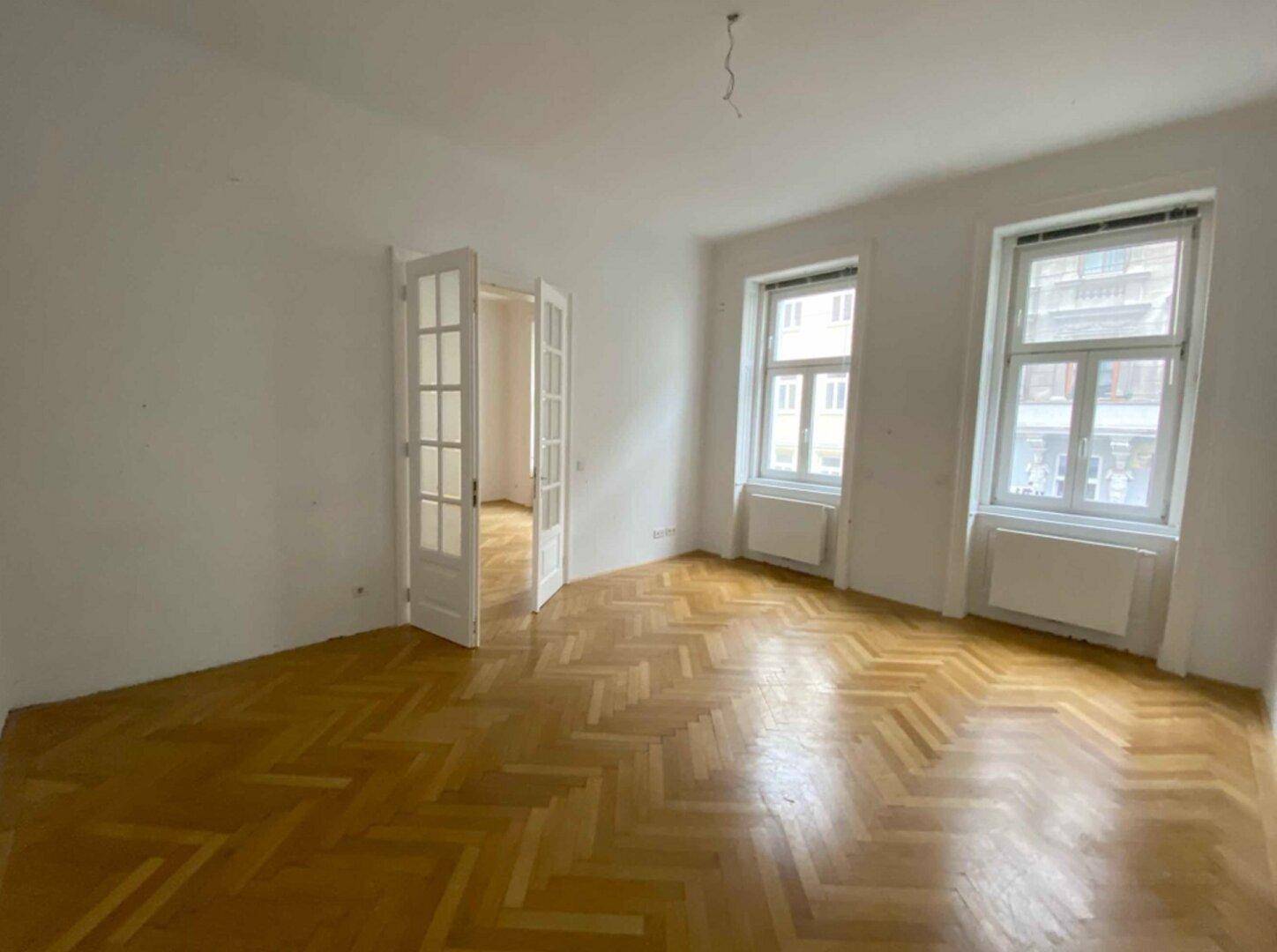 Leeres Wohnzimmer mit Fischgrätparkett, hohen Fenstern und Flügeltüren.
