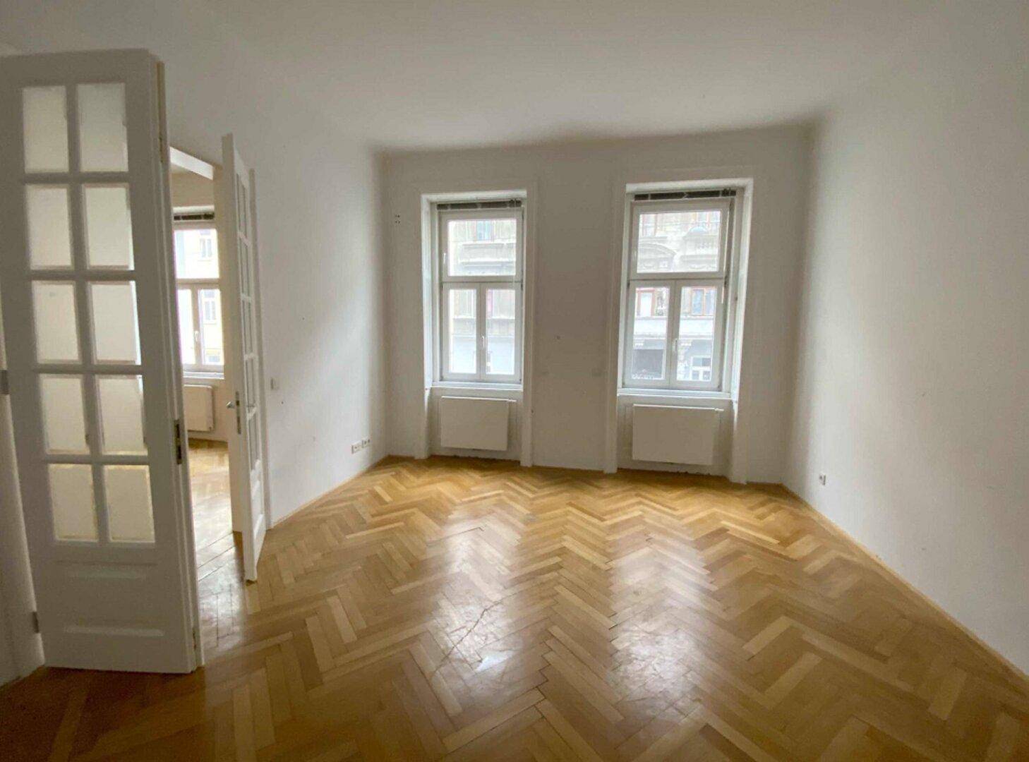 Heller Raum mit Fischgrätparkett und zwei großen Fenstern, die viel Tageslicht hereinlassen.
