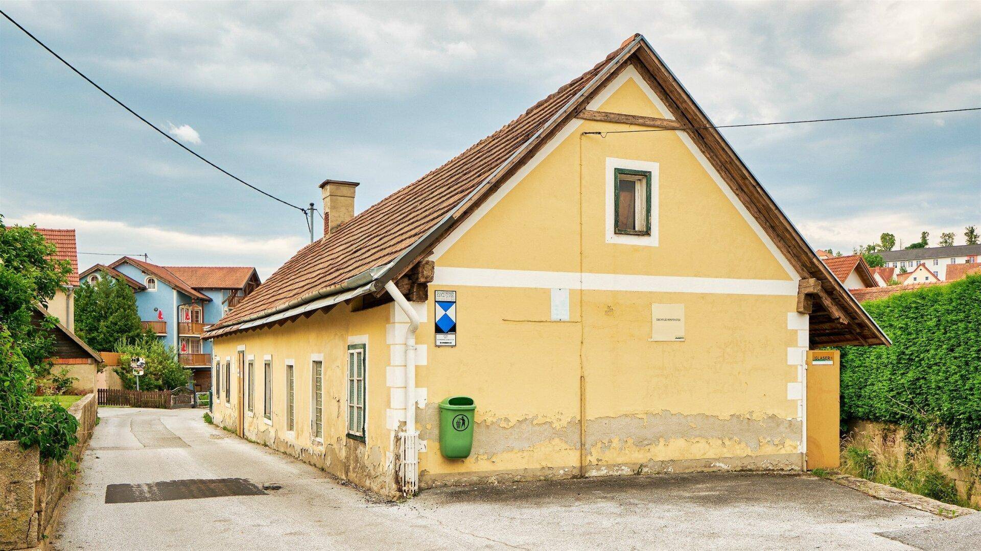 Seitenansicht eines gelben Gebäudes mit rotem Ziegeldach, einem Schornstein und einem grünen Mülleimer.
