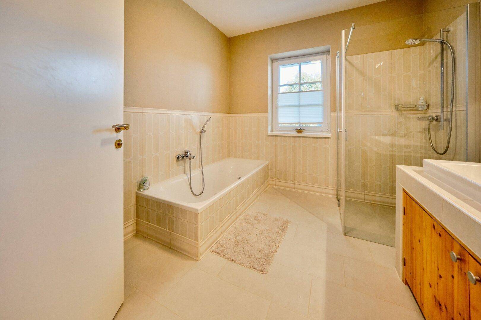 Geräumiges Badezimmer mit Badewanne, Duschkabine und Fenster, in hellen Farbtönen gehalten.