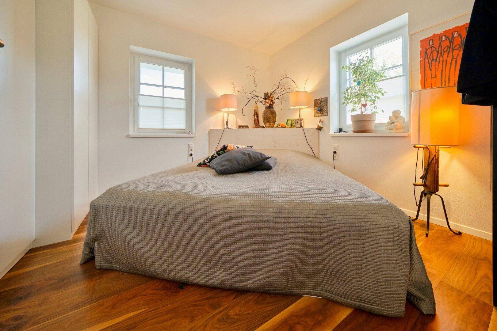 Gemütliches Schlafzimmer mit Holzboden, zwei Fenstern und indirekter Beleuchtung.