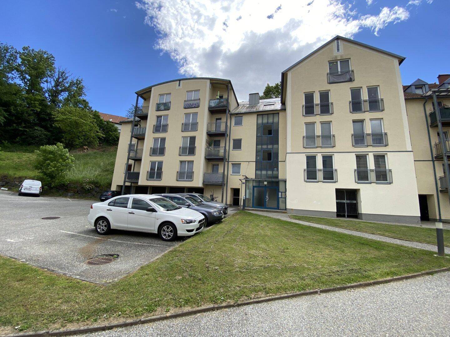 Ansicht des Apartmentgebäudes von der Seite mit Parkplätzen und einer grünen Hanglage im Hintergrund.