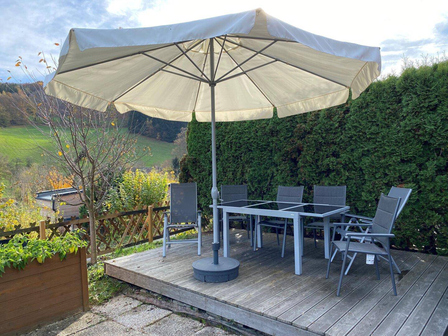 Gemütliche Holzterrasse mit Esstisch, Stühlen und Sonnenschirm, umgeben von Natur.