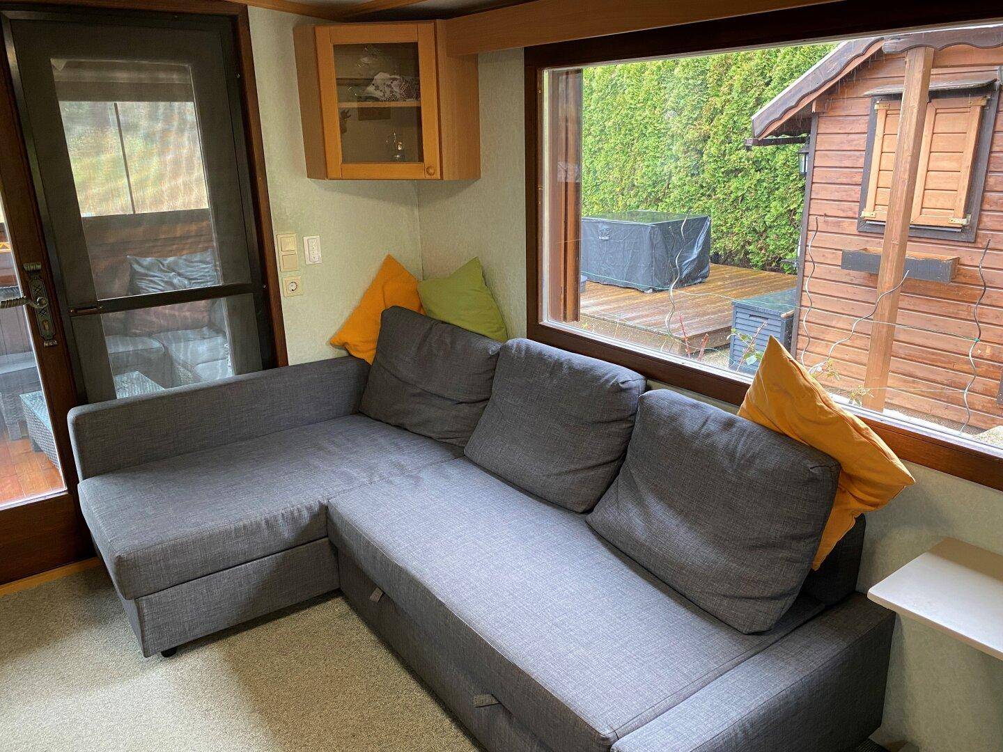 Helles Wohnzimmer mit grauem Ecksofa und Blick auf den Gartenbereich durch das Fenster.