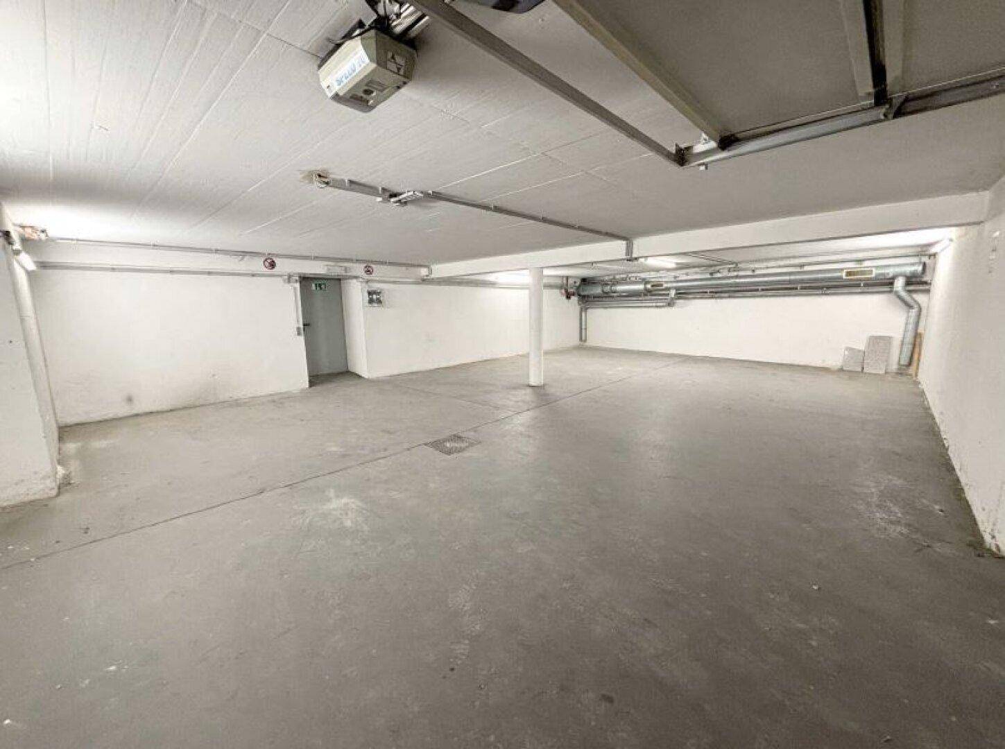 Großer Garagenbereich mit Betonboden, weißen Wänden und einem automatischen Toröffner an der Decke.
