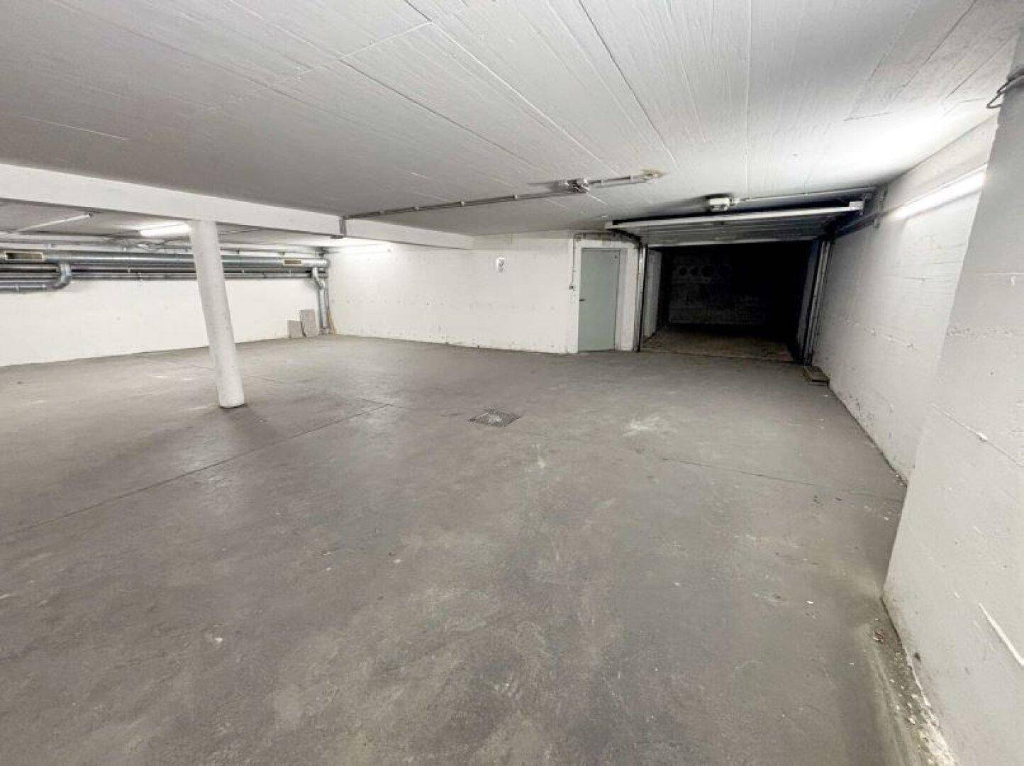 Geräumiger, leerer Garagenbereich mit Betonboden und sichtbaren Leitungen an der Decke.