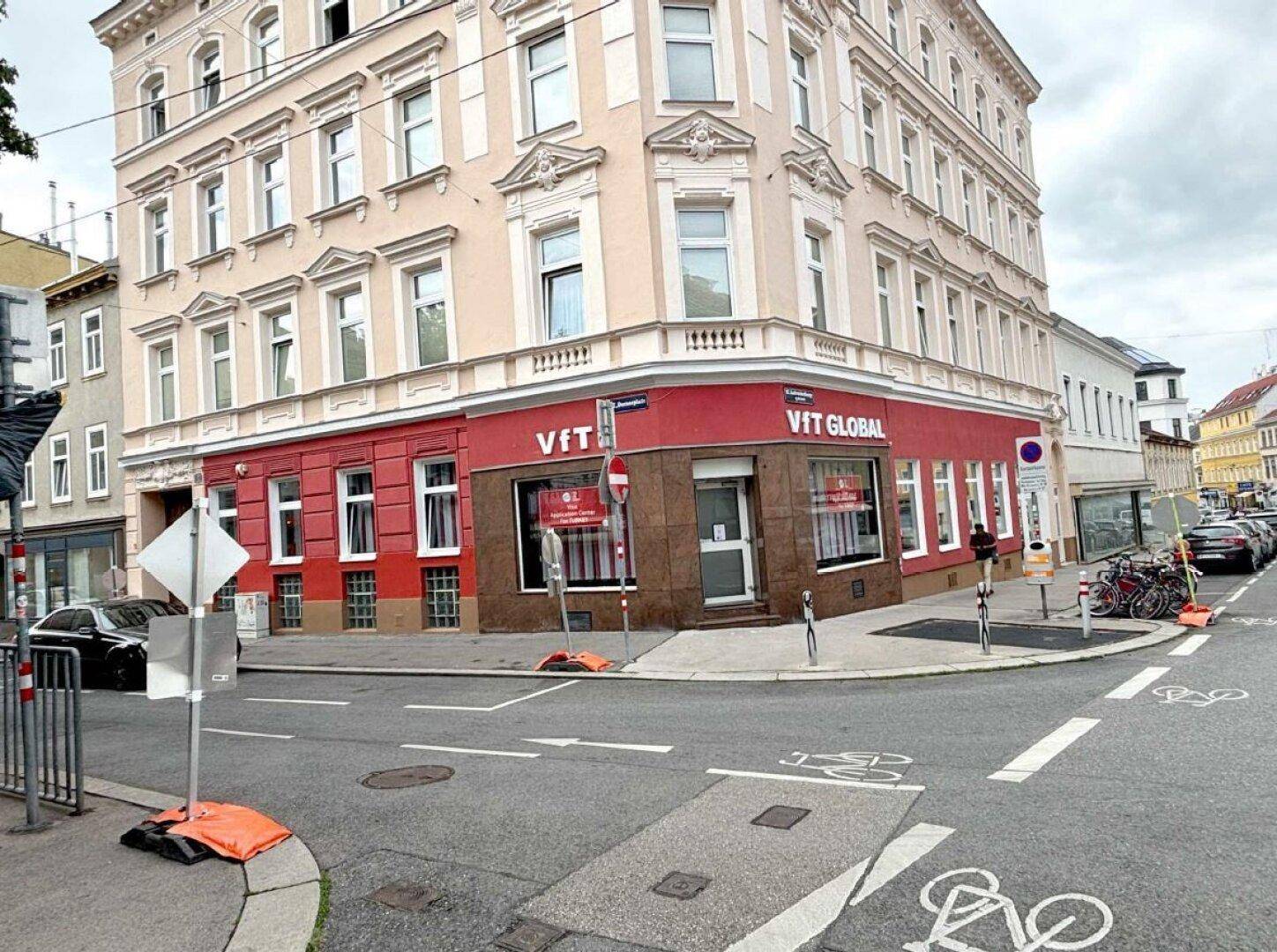 Eckgebäude mit roter Fassade im Erdgeschoss und hellen Obergeschossen an einer belebten Stadtstraße.