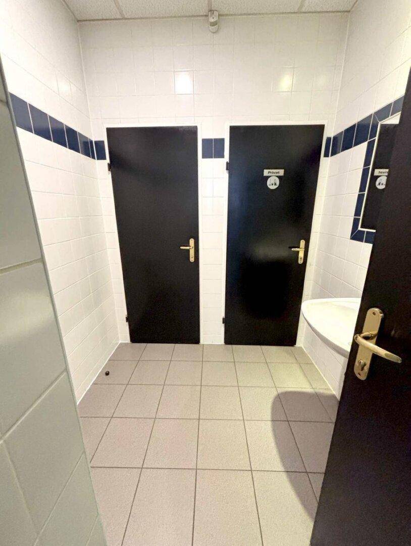 Sanitärbereich mit zwei schwarzen Türen, weißen Fliesenwänden und grauem Fliesenboden, sauber und funktional.