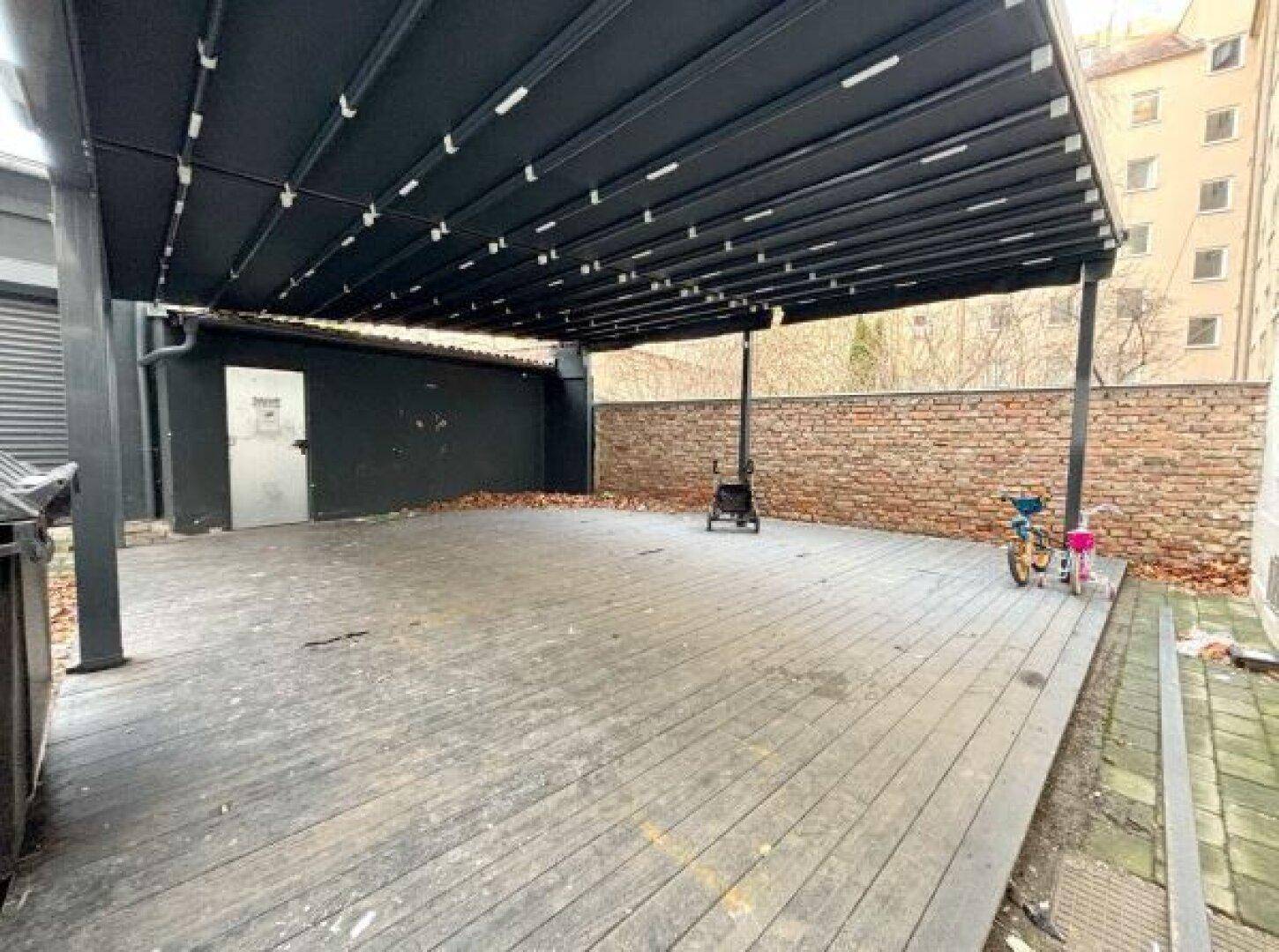 Große überdachte Holzterrasse mit dunklem Sonnenschutz und einer Ziegelmauer im Hintergrund.