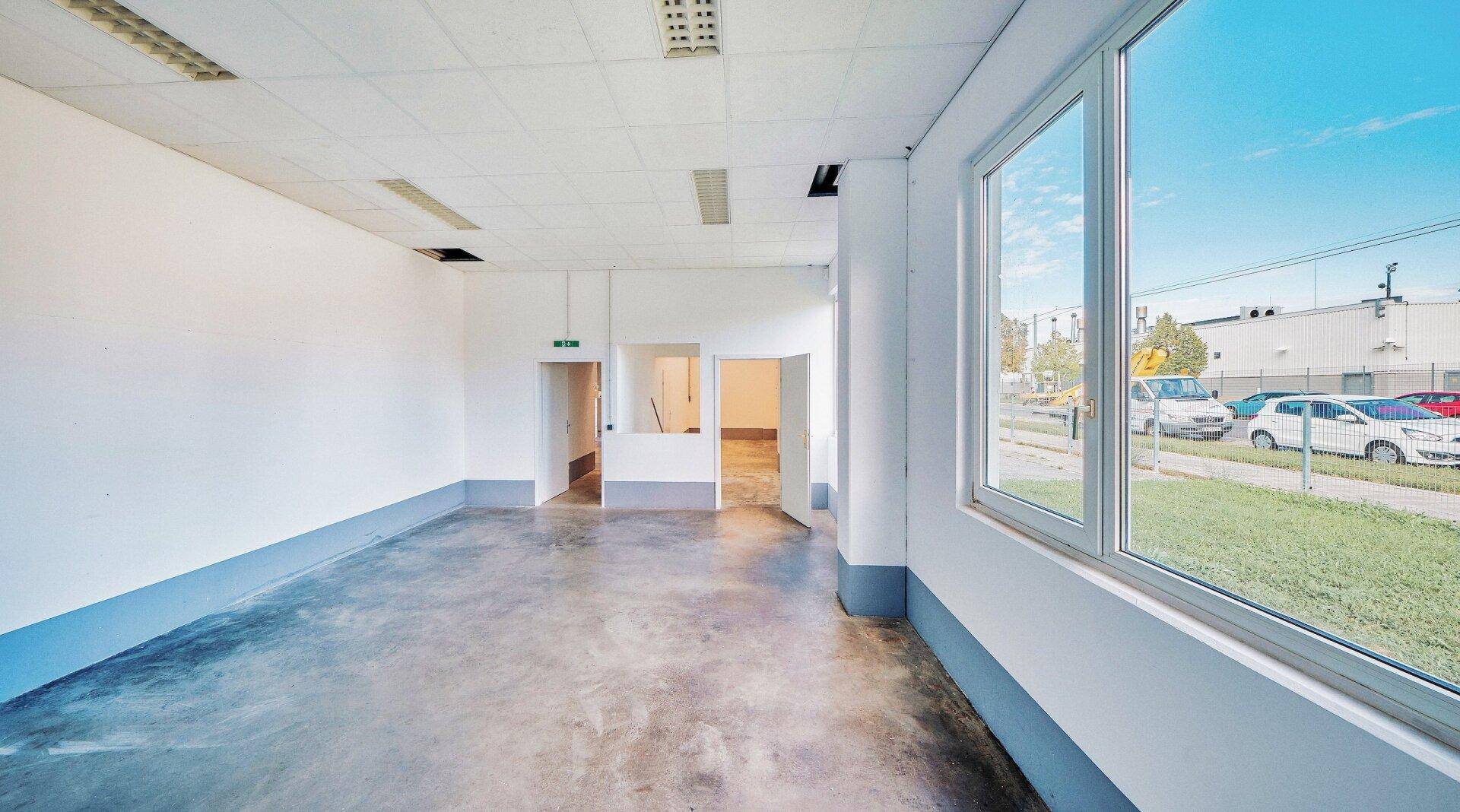 Heller Büroraum mit großen Fenstern, Betonboden und Blick auf den Außenbereich.