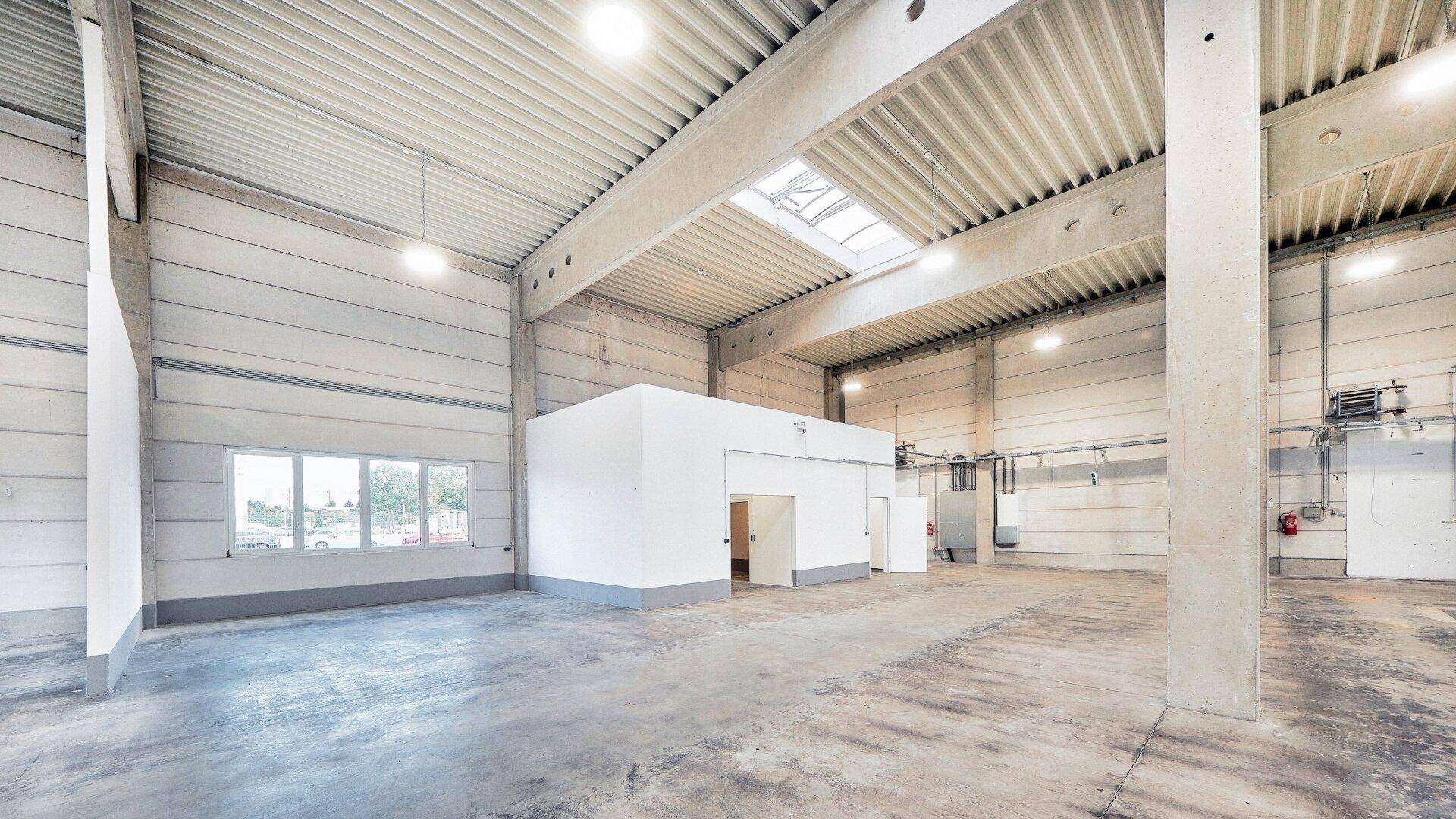 Geräumige Lagerhalle mit hohen Decken, Betonboden und einem abgetrennten Bürobereich.
