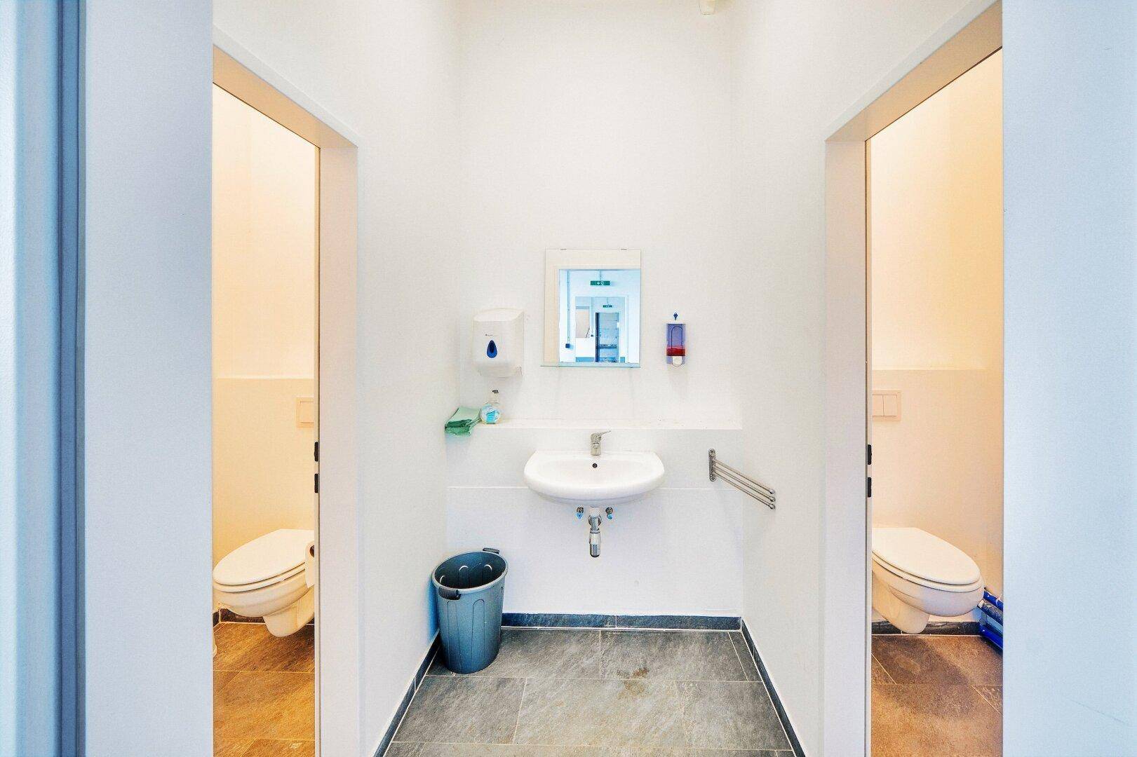 Zwei separate Toilettenräume mit Waschbecken, Spiegel und Fliesenboden in einem Vorraum.