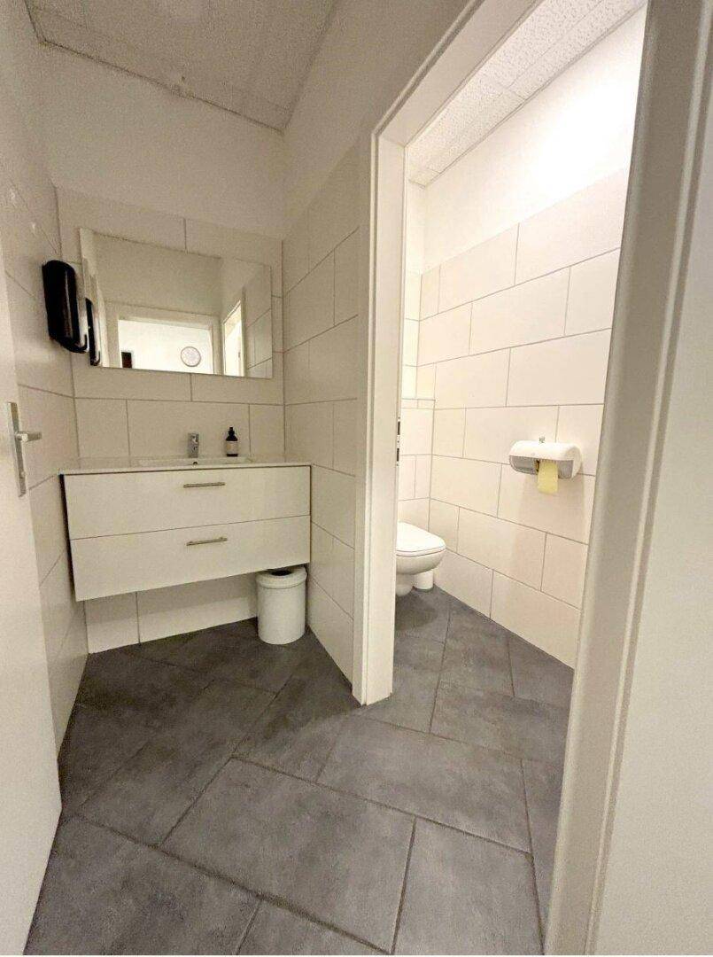 Modern gefliestes WC mit Hänge-Waschtisch, Spiegel und dunklem Boden in einem hellen Raum.