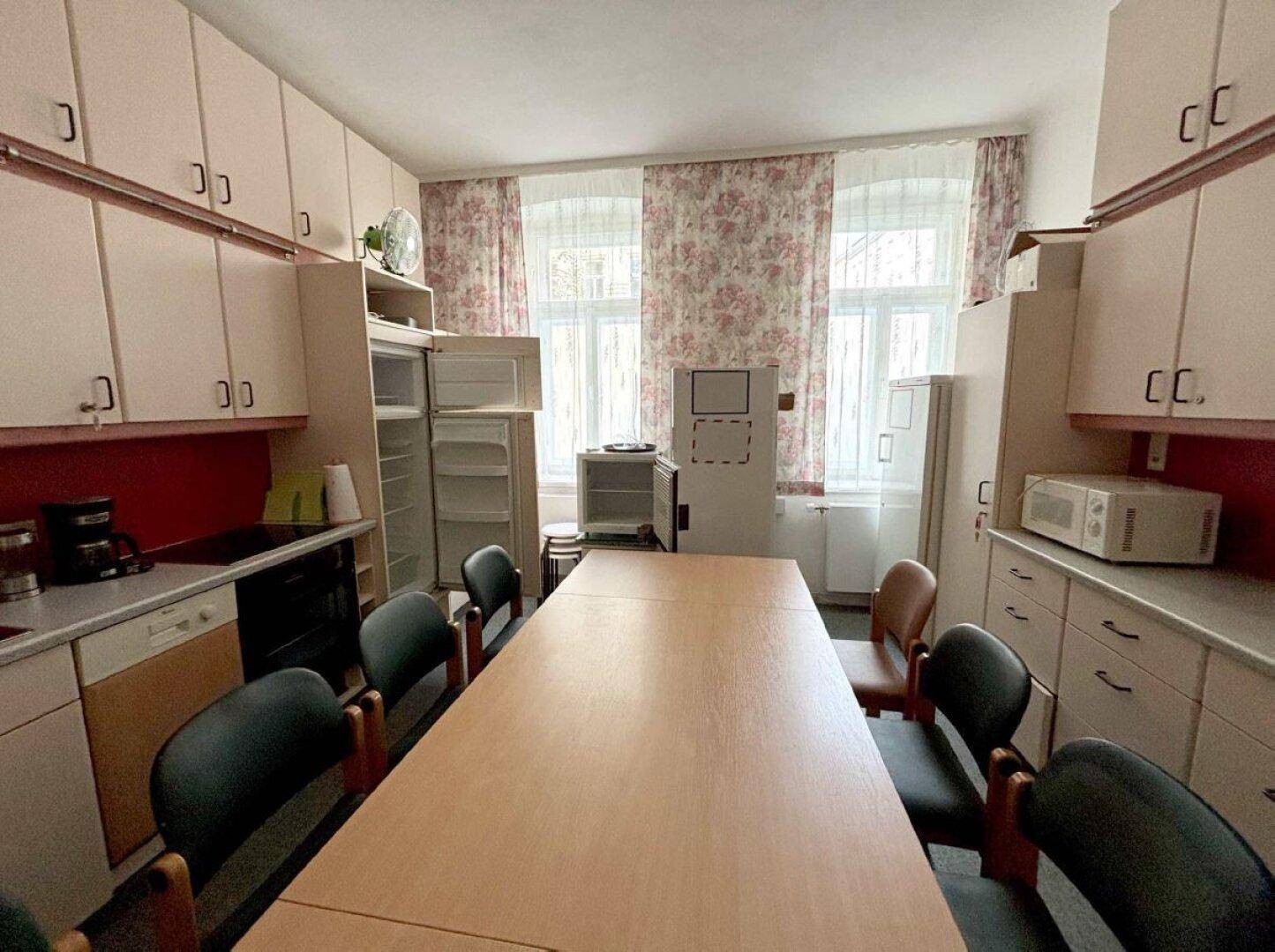Heller Büro- oder Pausenraum mit großer Tafel, Küchenzeile und Fenstern mit floralen Vorhängen.