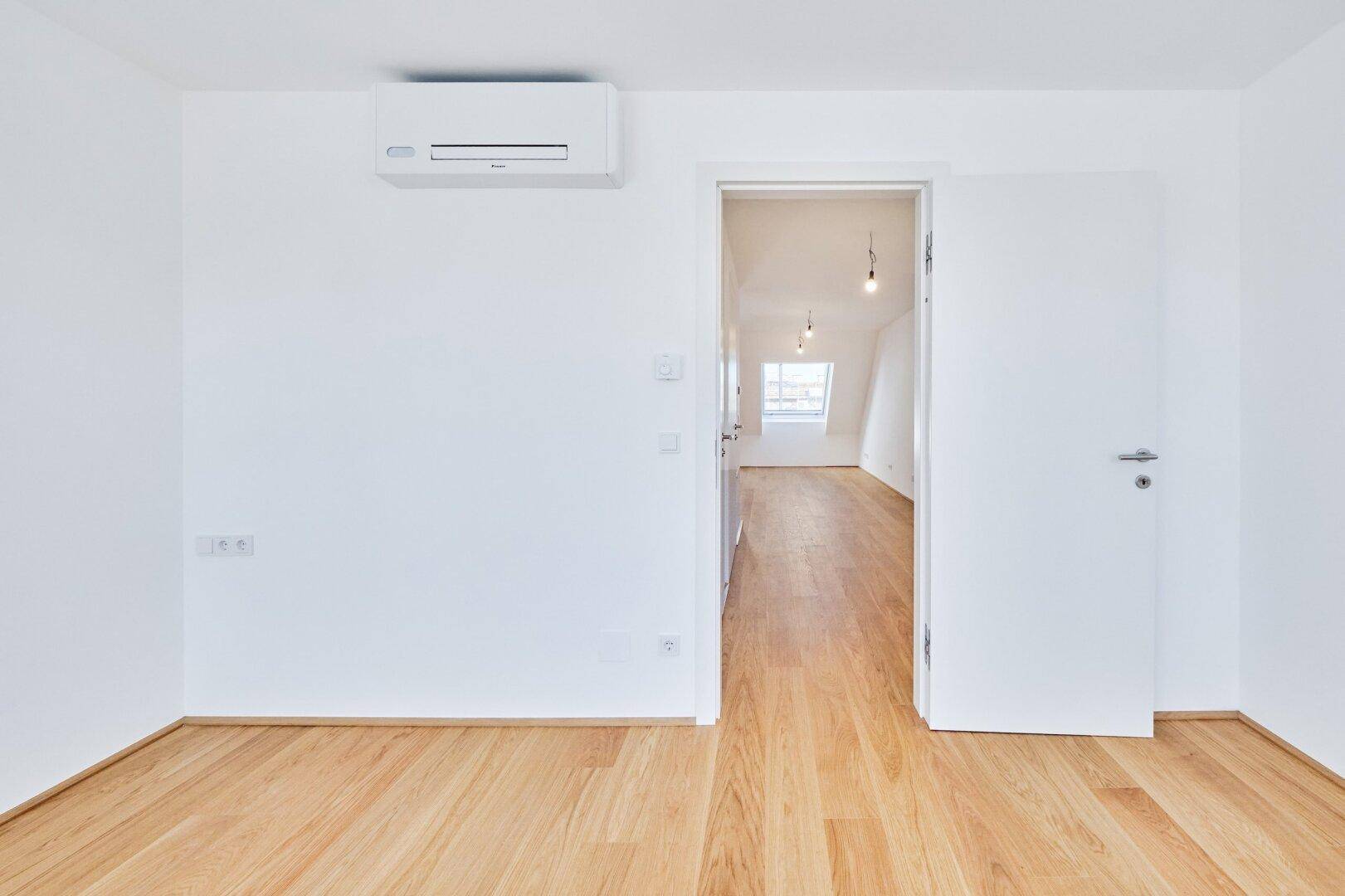 Leerer Raum mit Holzboden, weißen Wänden und Klimaanlage, offen zu einem weiteren Zimmer.