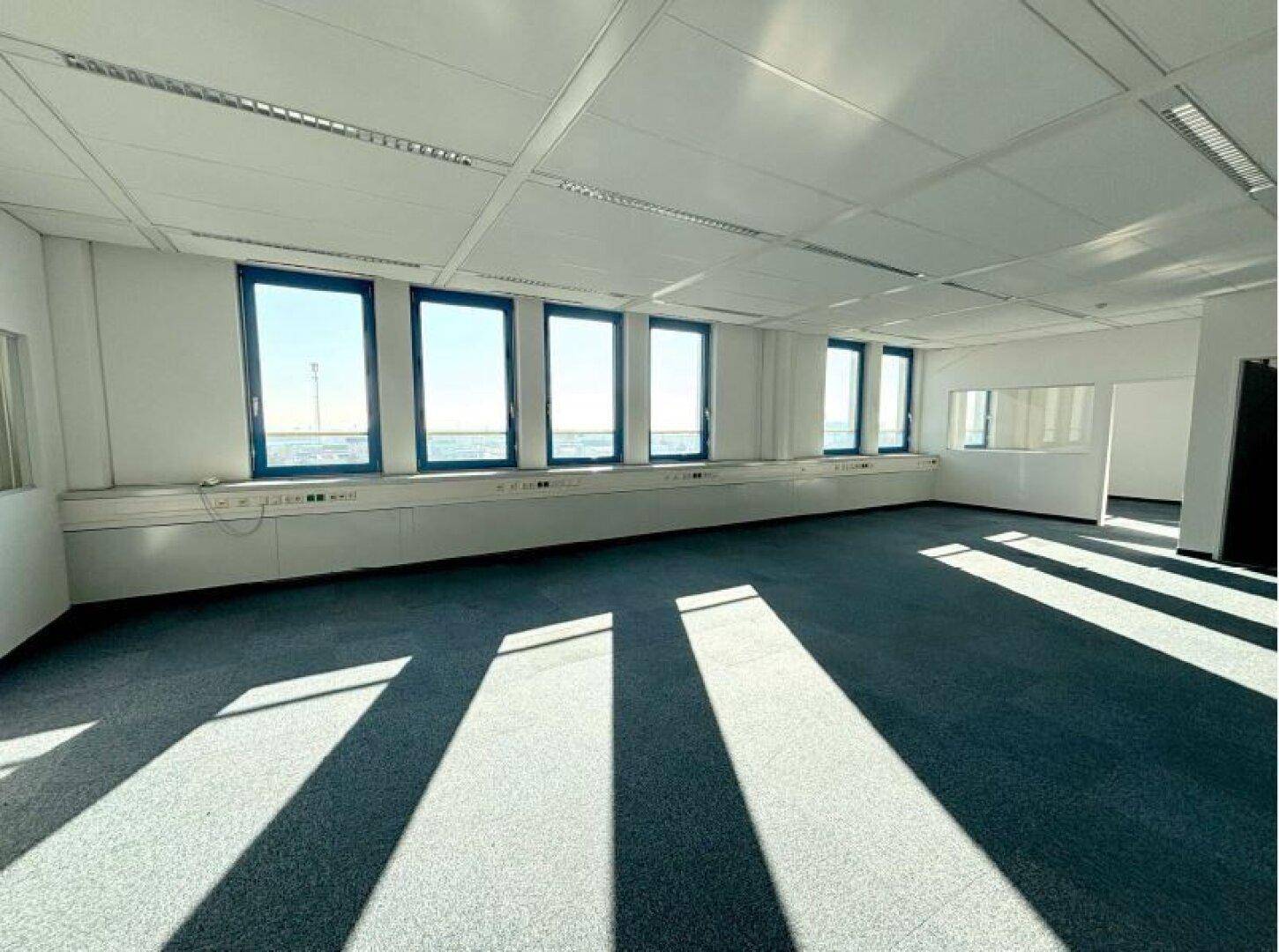 Geräumiges, leeres Büro mit Teppichboden und großen Fenstern, die viel Tageslicht hereinlassen.