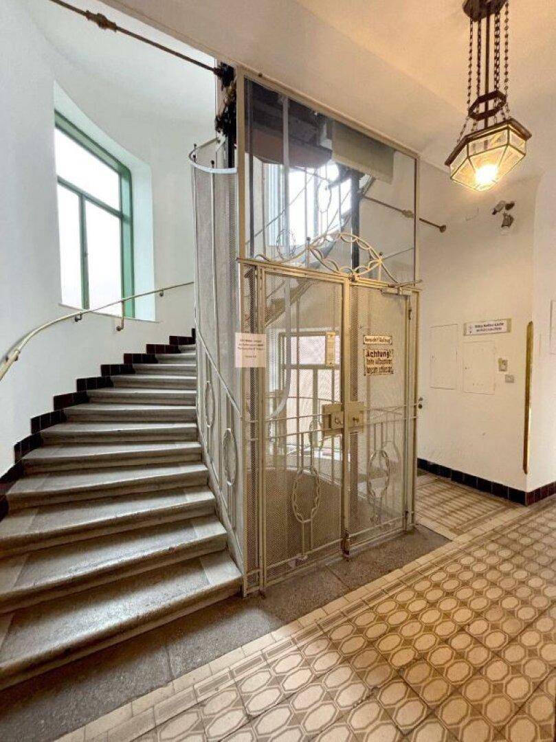 Historisches Treppenhaus mit elegantem Aufzug, geschwungener Treppe und dekorativer Beleuchtung.