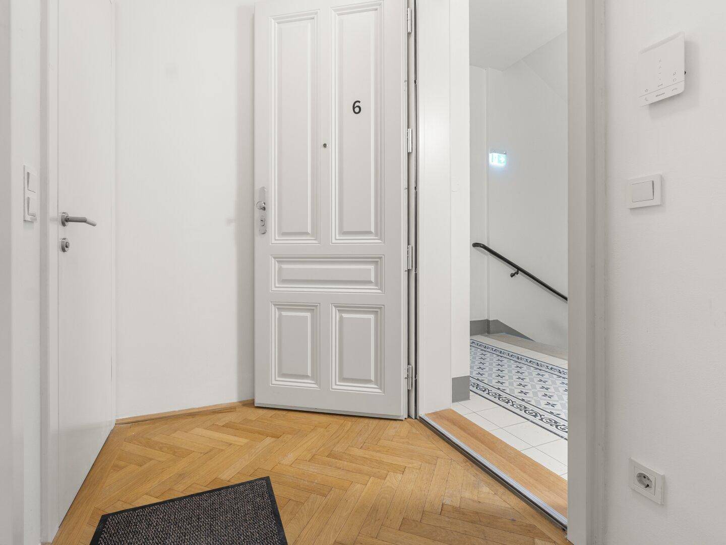 Heller Flur mit Fischgrätparkett und weißer Tür, die zum Treppenhaus mit gemusterten Fliesen führt.