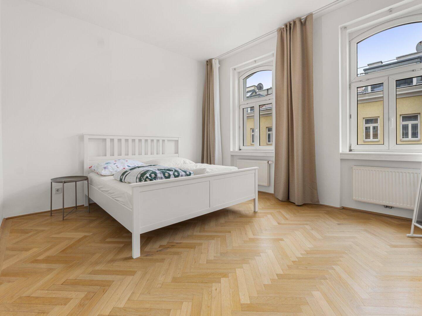 Geräumiges Schlafzimmer mit klassischem Fischgrätparkett und zwei hohen Fenstern.