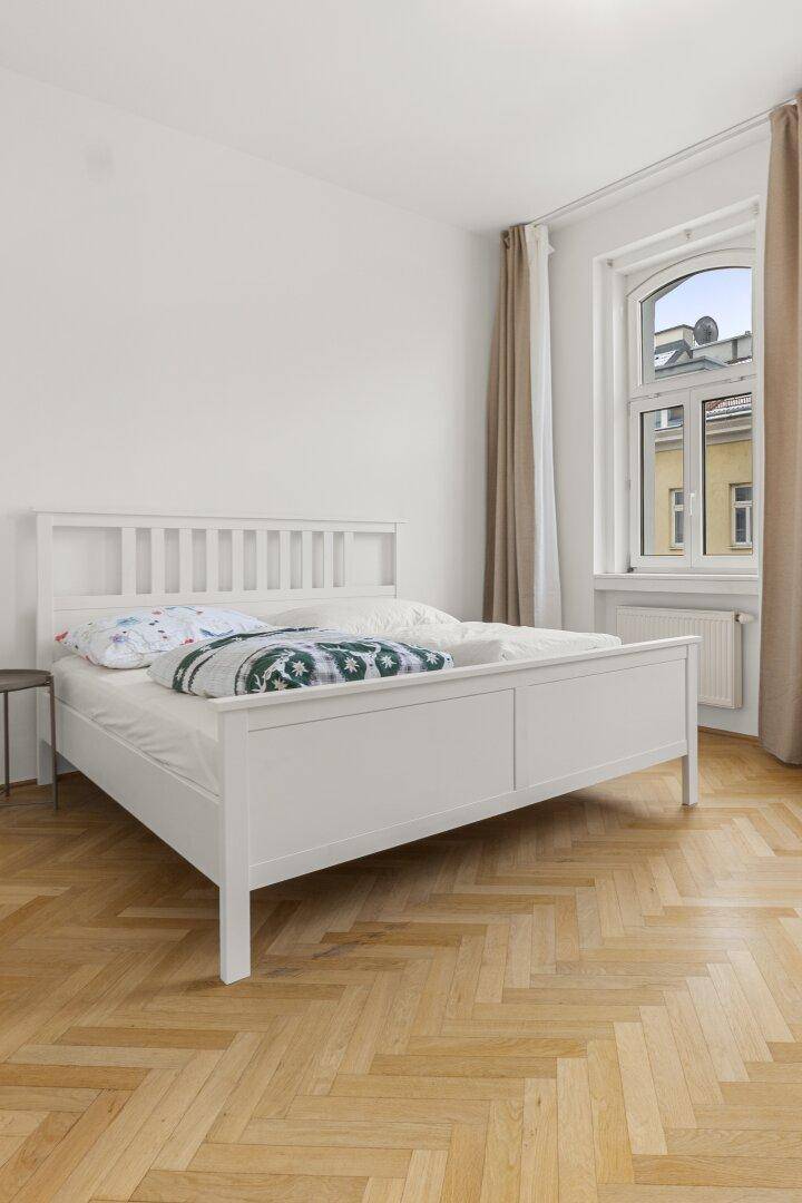 Schlafzimmer mit weißem Bett, hellem Fischgrätparkett und großem Fenster für viel Tageslicht.