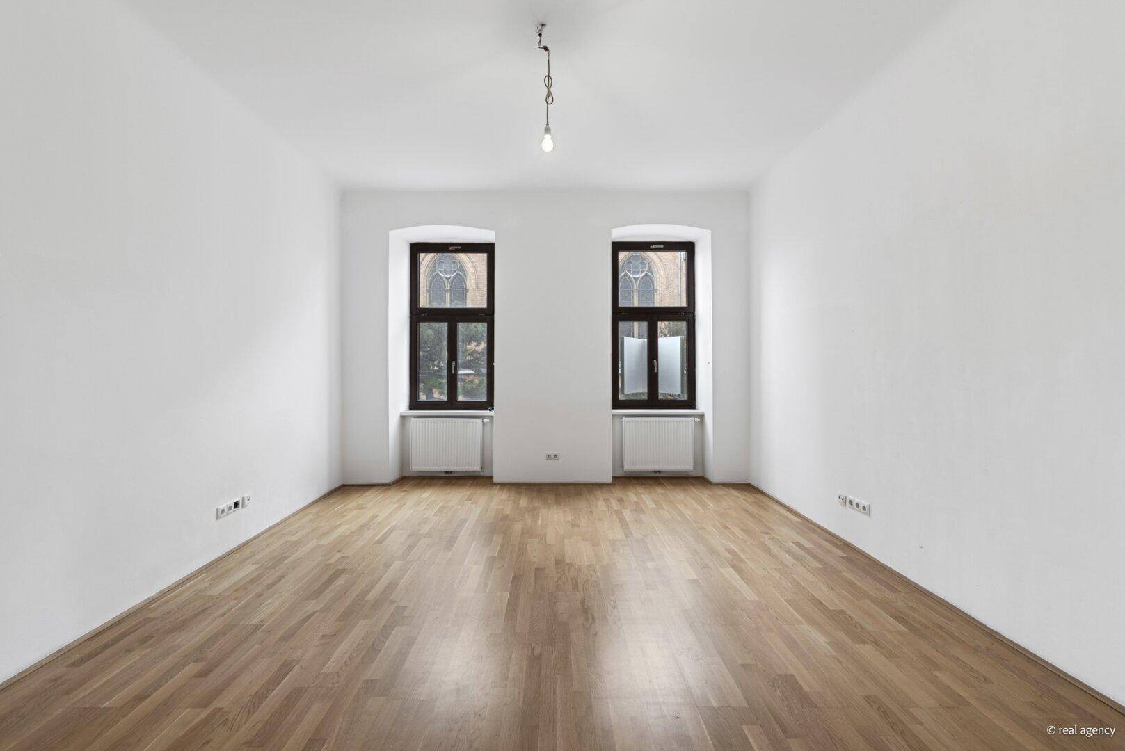 Geräumiges, leeres Zimmer mit Parkettboden und zwei hohen Fenstern, bietet viel Gestaltungspotenzial.
