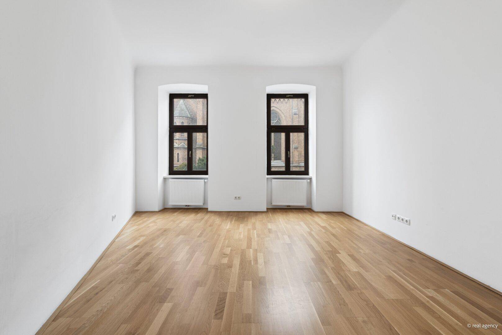 Heller Raum mit Parkettboden und zwei Fenstern, die einen Blick nach draußen ermöglichen.
