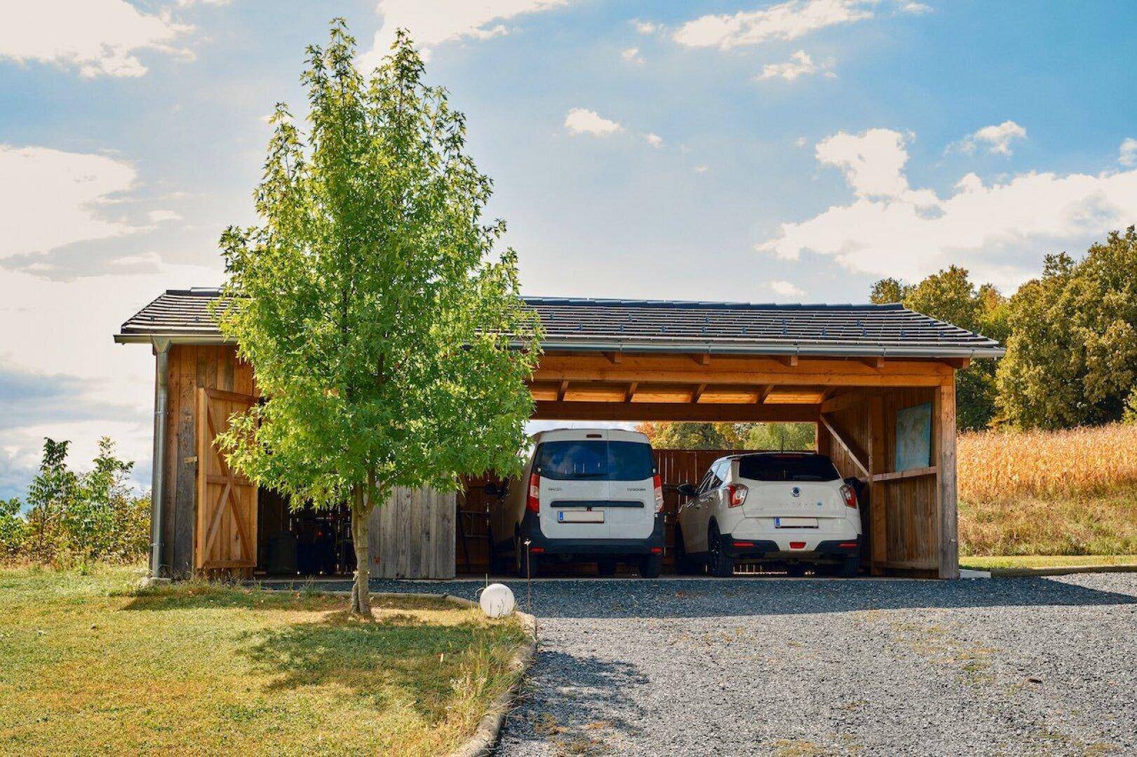 Offener Holzcarport mit zwei Stellplätzen für Fahrzeuge, umgeben von grüner Landschaft.