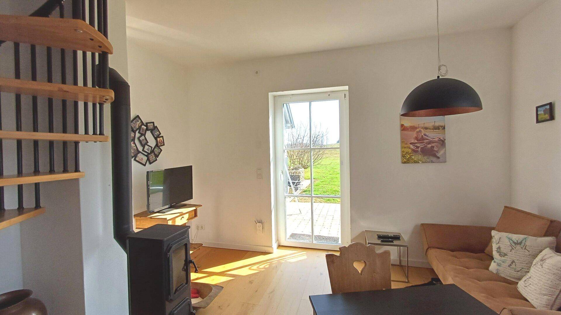 Helles Wohnzimmer mit Holzofen, Wendeltreppe und Zugang zur Terrasse mit Grünblick.
