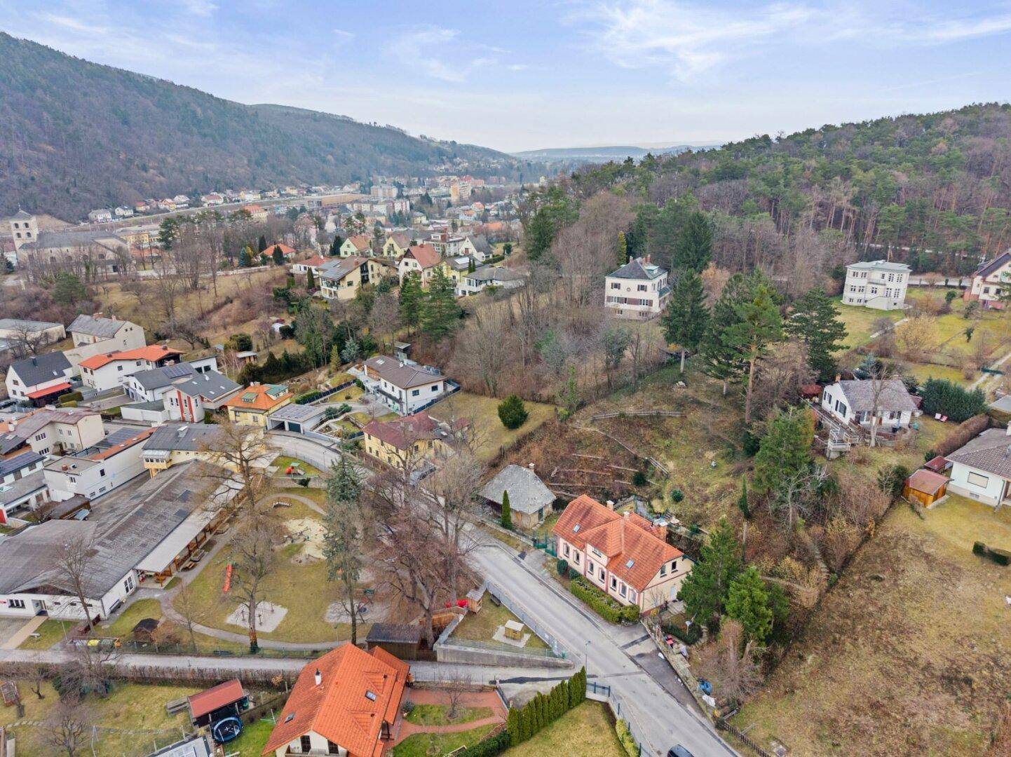 Luftaufnahme einer Siedlung in einem Tal, umgeben von bewaldeten Hügeln und Wohnhäusern.