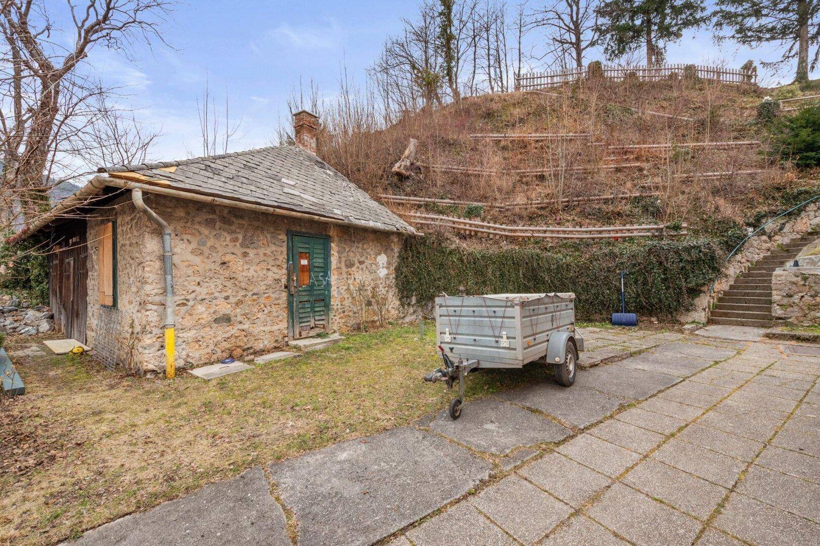 Kleines Nebengebäude aus Stein mit gepflastertem Vorplatz und einem Anhänger.
