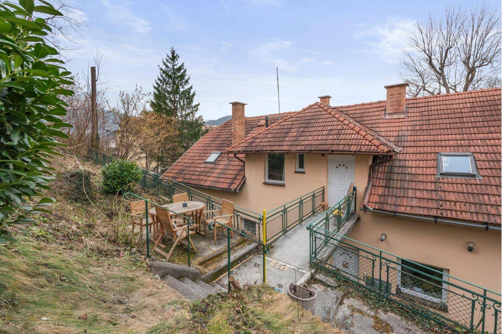 Kleiner Außenbereich mit Holztisch und Stühlen neben dem Haus, umgeben von Hanglage.