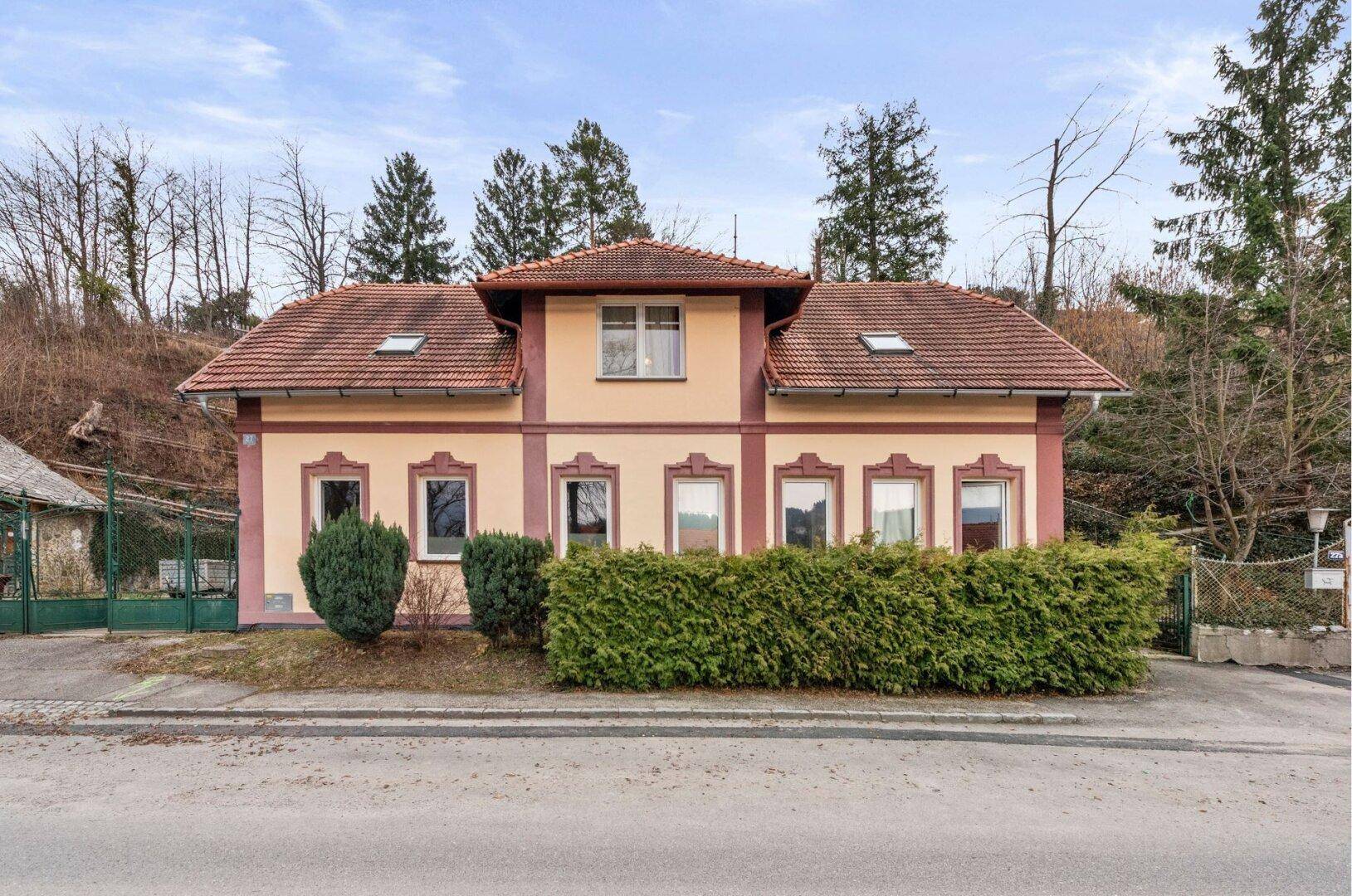 Fassade eines traditionellen Hauses mit hellbraunem Putz, roten Ziegeldächern und grüner Hecke.