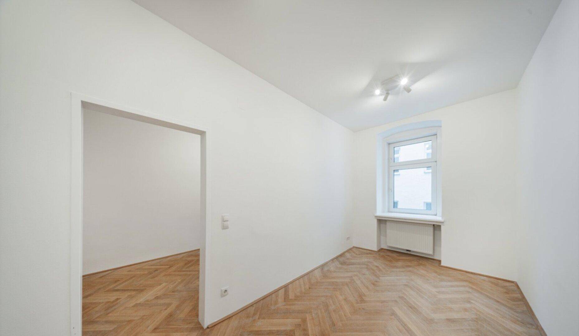 Helles Zimmer mit Fischgrätparkett, weißen Wänden und einem Fenster mit Ausblick.