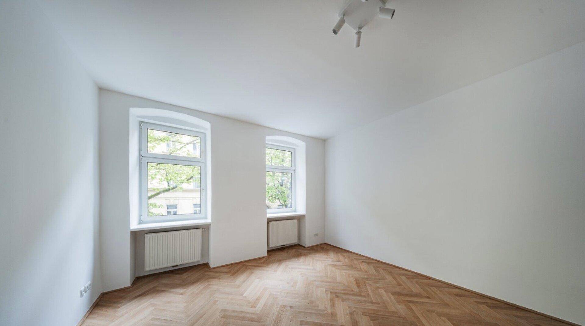 Geräumiges Zimmer mit Fischgrätparkett und zwei Fenstern, die viel Licht hereinlassen.