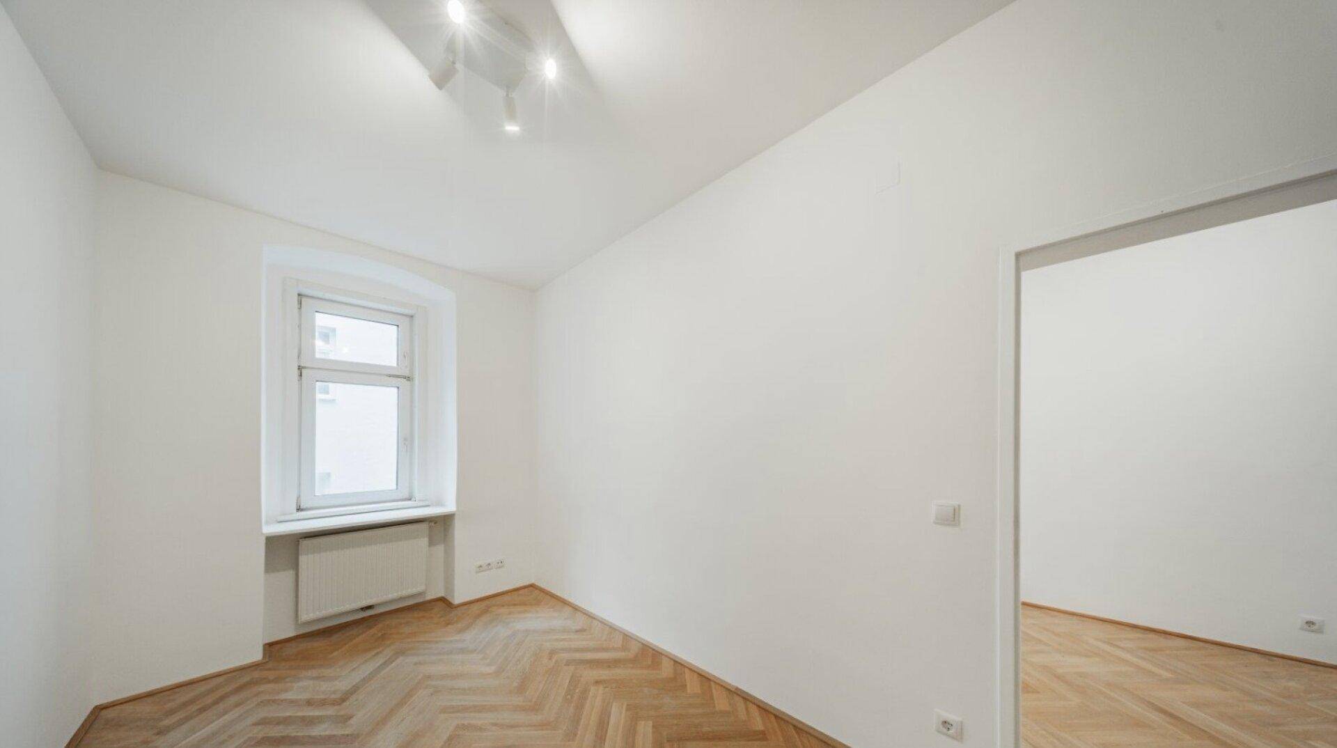 Helles Zimmer mit Fischgrätparkett, weißen Wänden und einem Fenster.