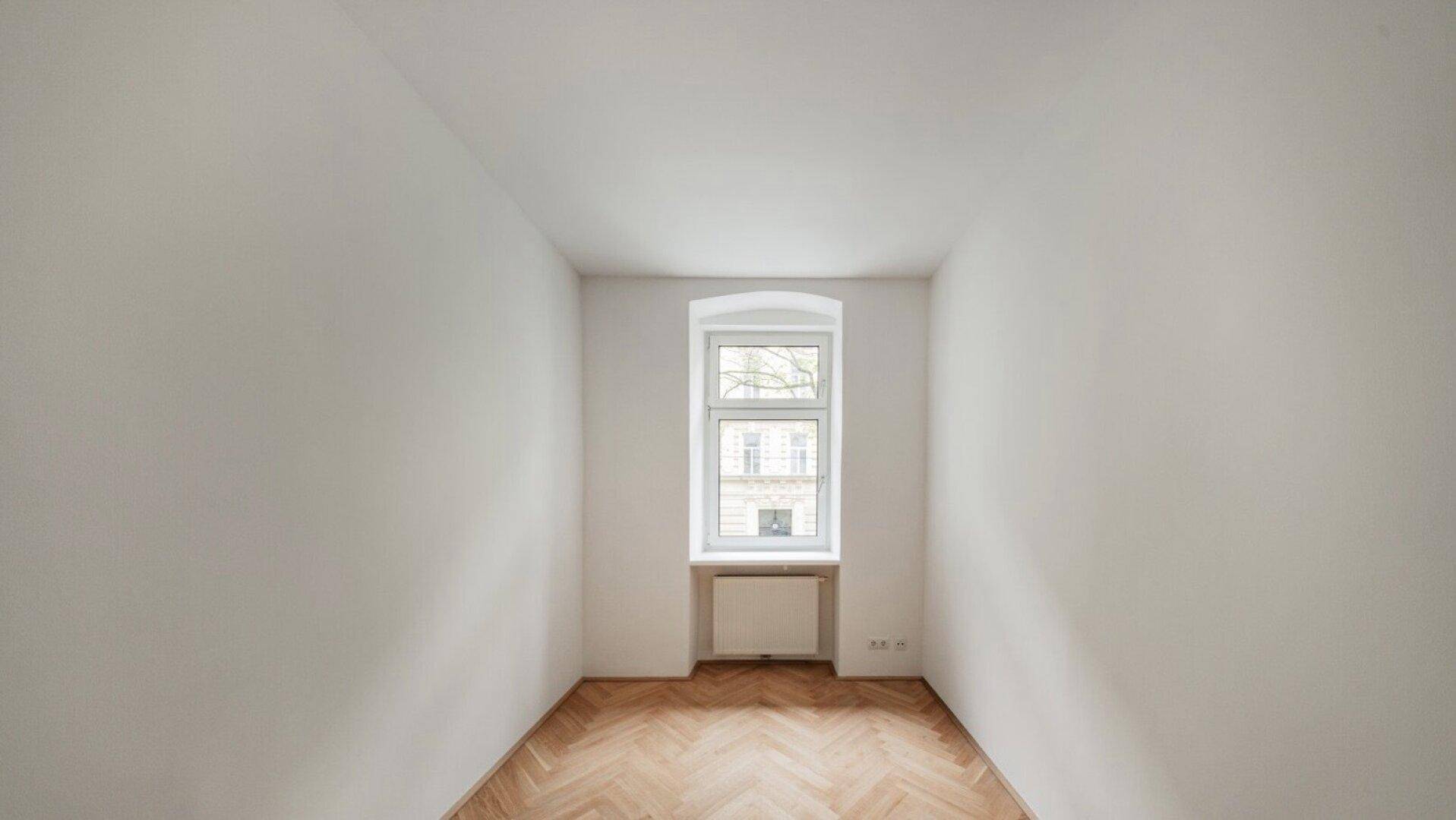 Schlichtes Zimmer mit Fischgrätparkett und einem Fenster, das Tageslicht spendet.