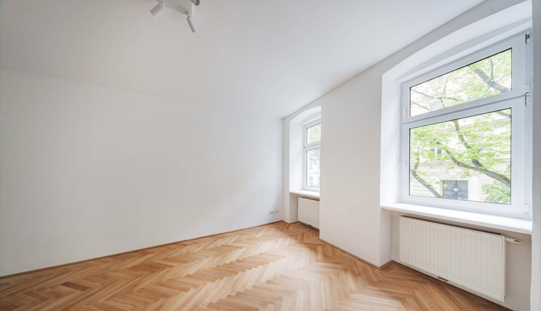 Geräumiges Zimmer mit hellem Fischgrätparkett und zwei Fenstern, die viel Tageslicht hereinlassen.